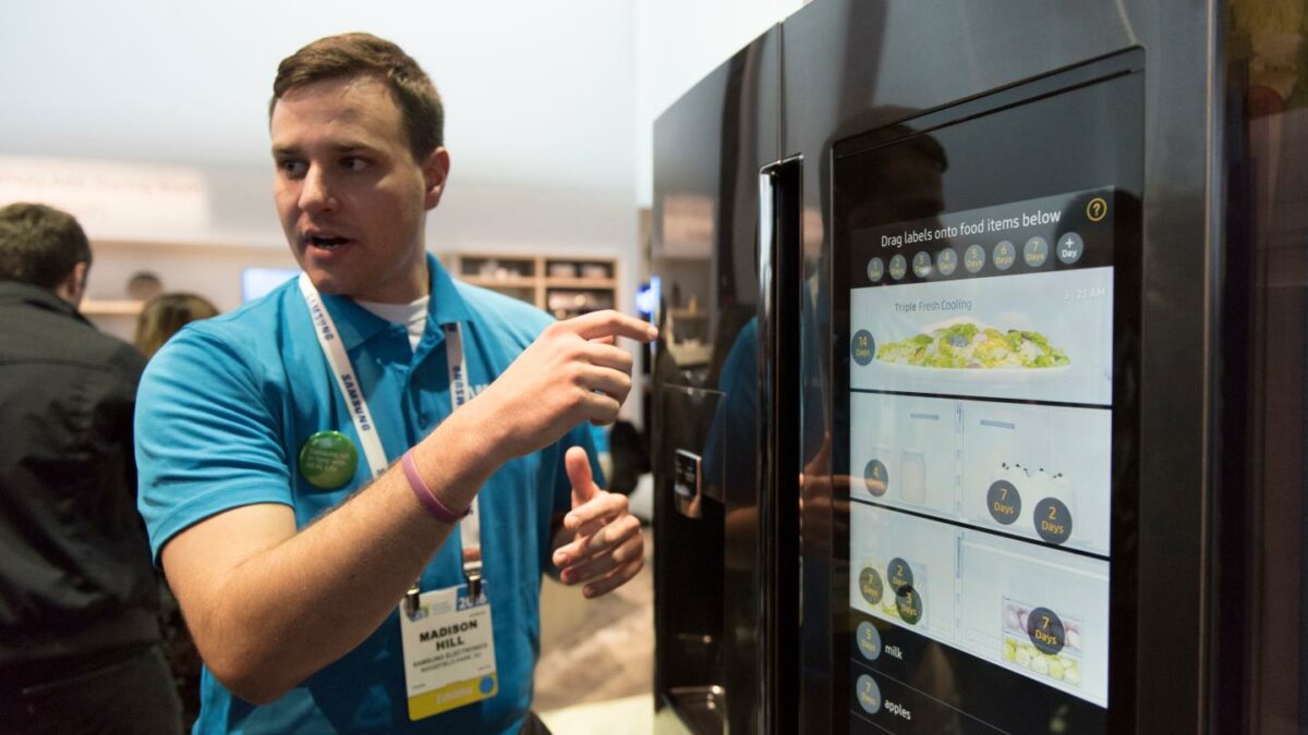 A man in a blue short points to the display screen on a Samsung refrigerator.