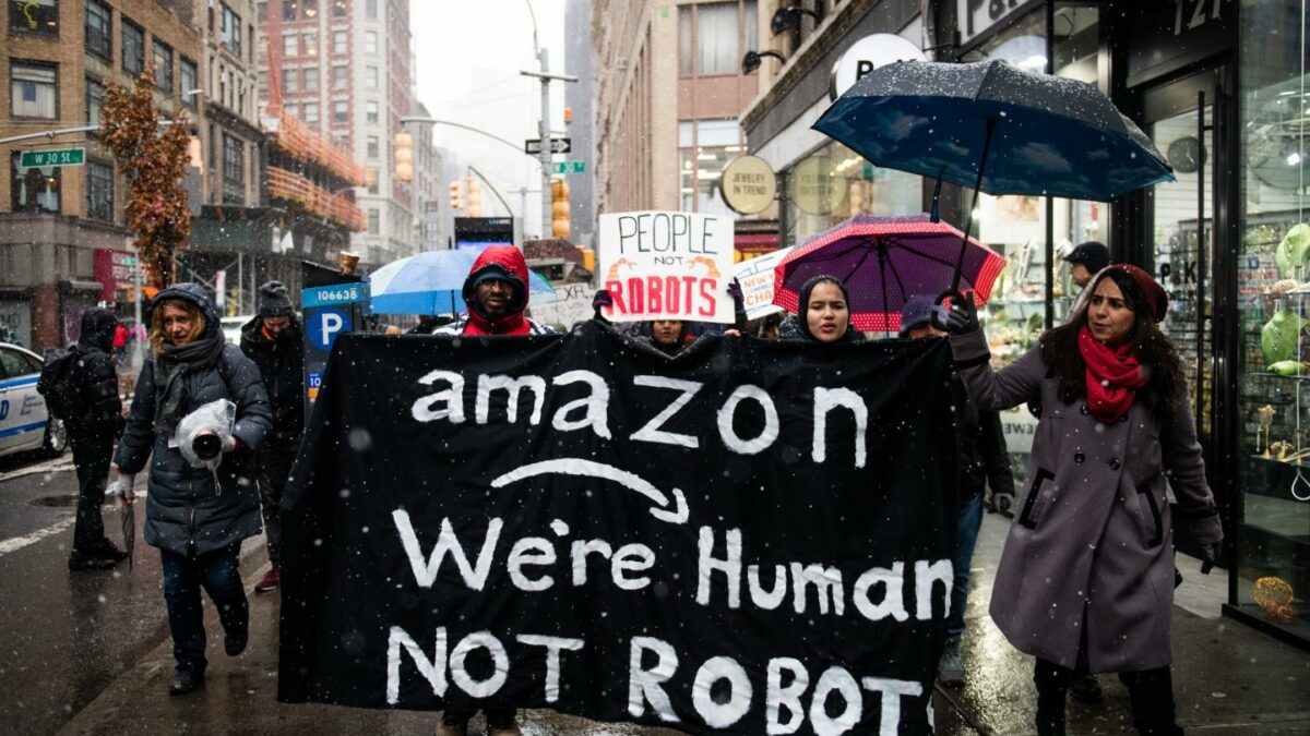 Amazon workers hold a large black sign that reads, "Amazon, We're humans, Not robots."