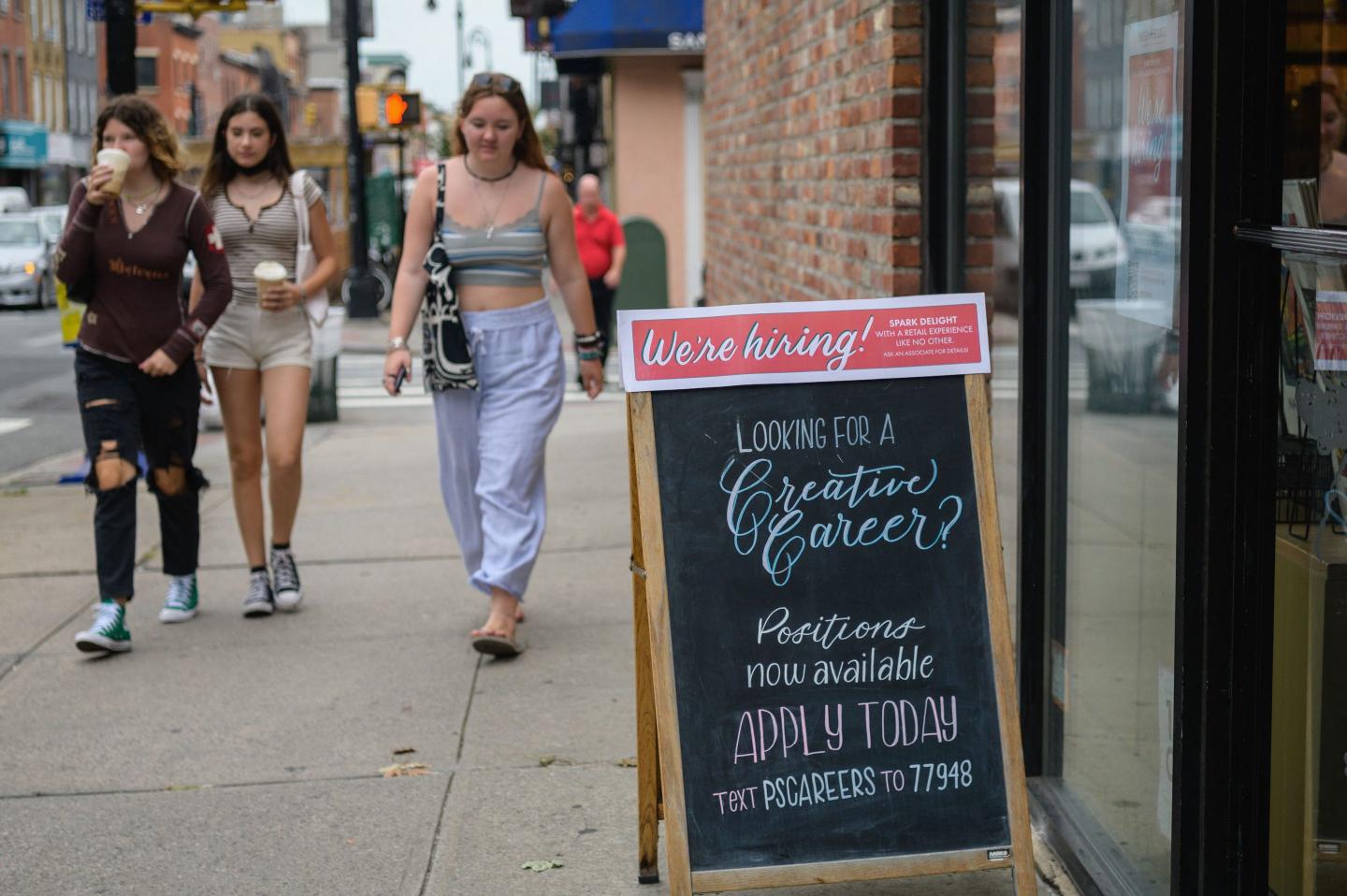 A group of young people walk past a sign that says We’re hiring!