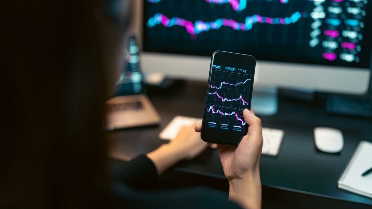 A woman trades on her phone in front of a computer with a display of crypto charts.