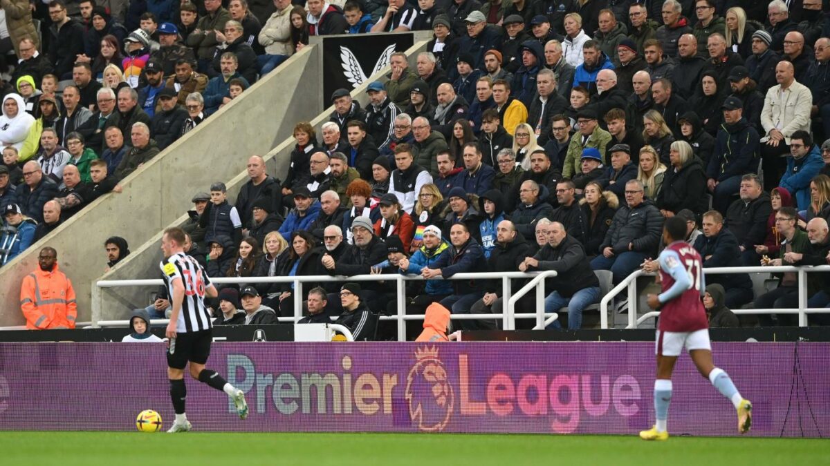 Two soccer players run across the field in front of bleachers filled with fans and a Premier League advertisement.