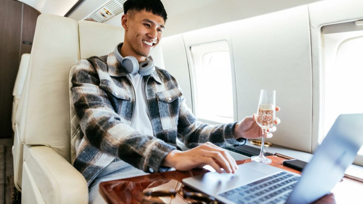 Young man sitting on a private jet, wearing a flannel while working on a laptop and holding a glass of champagne