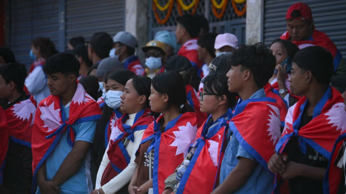 A group of young people stand together, each with the flag of Nepal draped around their shoulders.