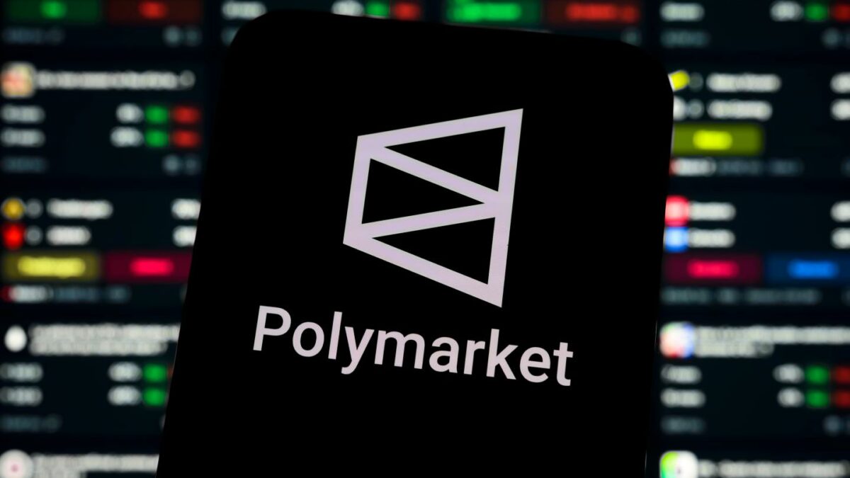 The logo of the decentralized prediction market platform Polymarket displays on the screen of a smartphone placed in front of a background showing a list of active bets,