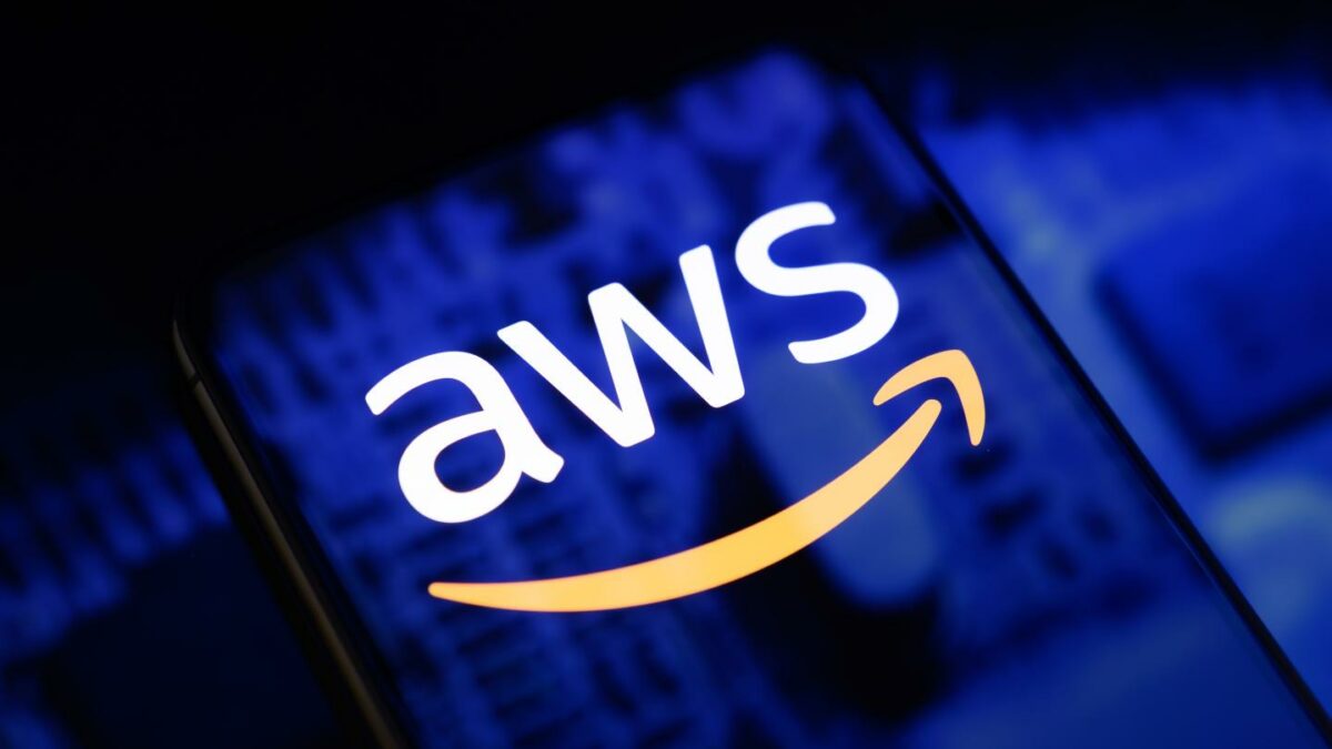 The AWS logo is displayed on a smartphone screen placed on a reflective surface onto which an illustration of blue printed circuit boards.