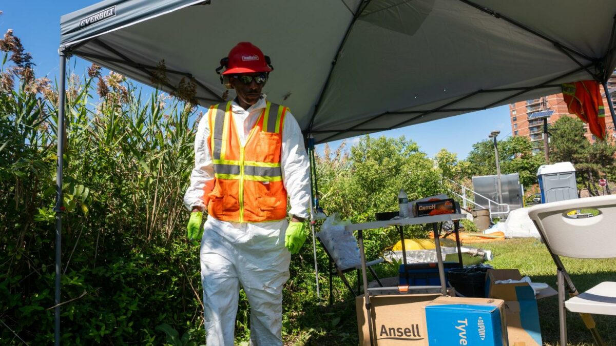 A man in an orange vest stands under a tent by a creek.