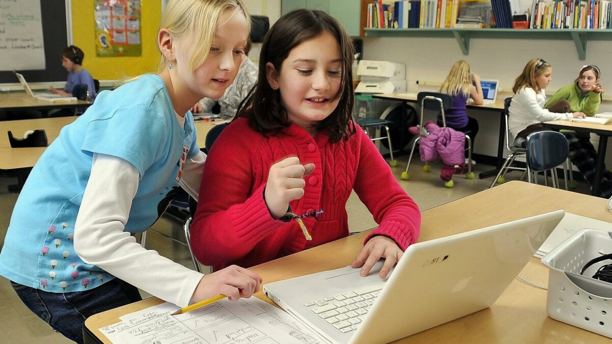 Two girls look at a laptop placed on a desk.