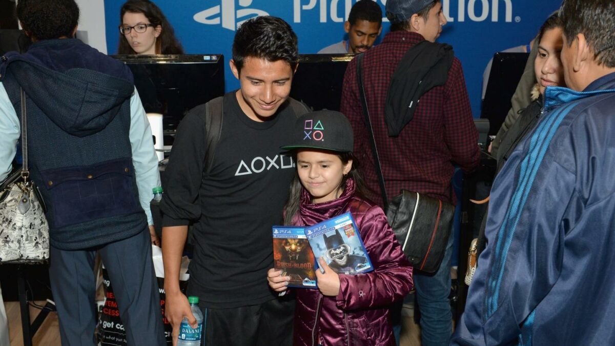A child holds up two video game cases and stands next to an adult wearing a PlayStation shirt.
