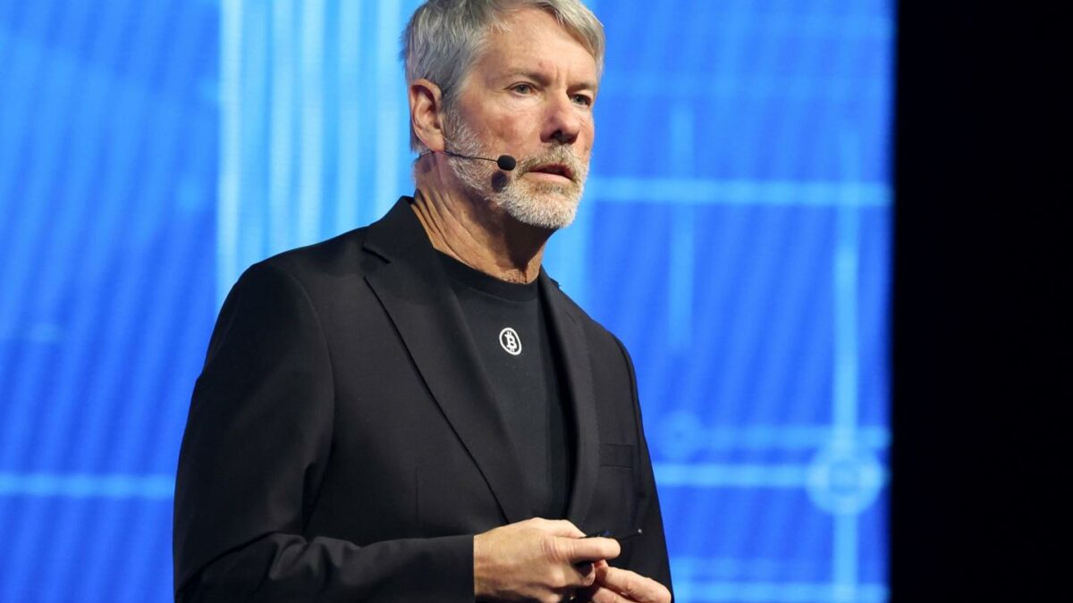Michael Saylor with a cheek microphone, wearing a black suit, and wearing a Bitcoin pin.